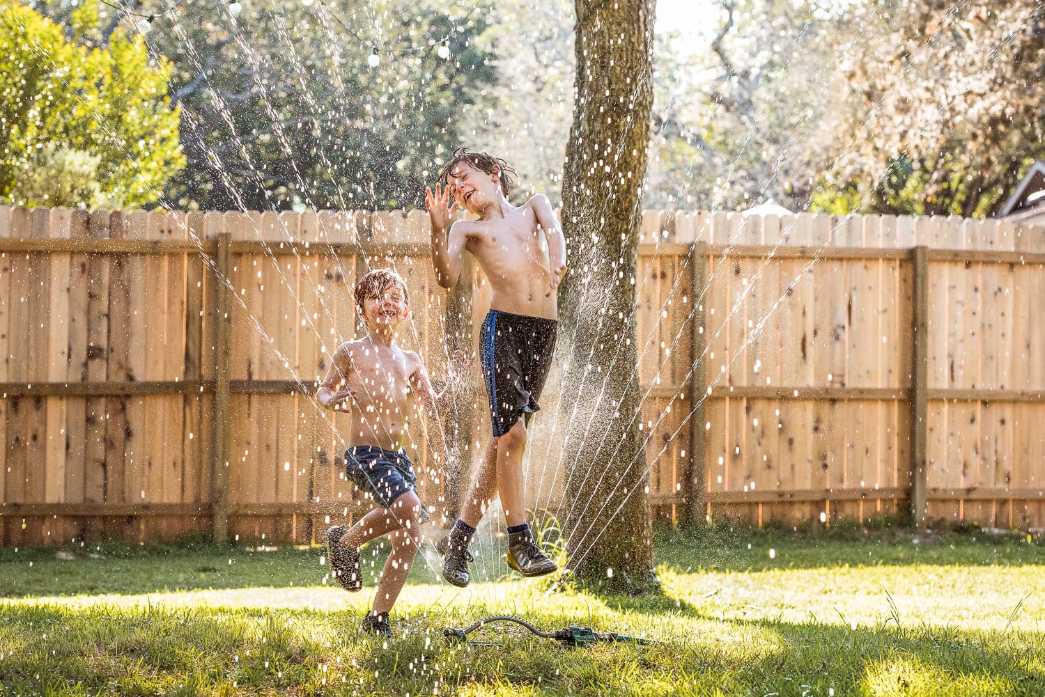 Two young boys jumping joyously through a backyard water sprinkler.